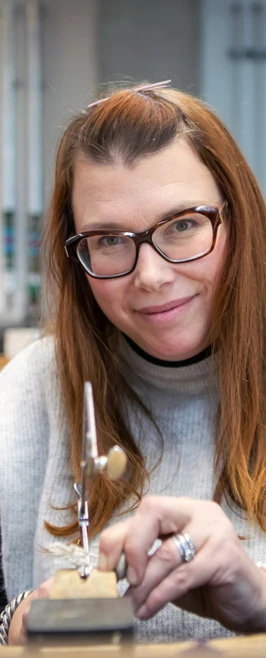 A female jewellery maker with long auburn hair and glasses, wearing a grey sweater, sitting at a workbench and looking directly at the camera while working on a piece of jewellery. A female jewellery maker with long auburn hair and glasses, wearing a grey sweater, sitting at a workbench and looking directly at the camera while working on a piece of jewellery.